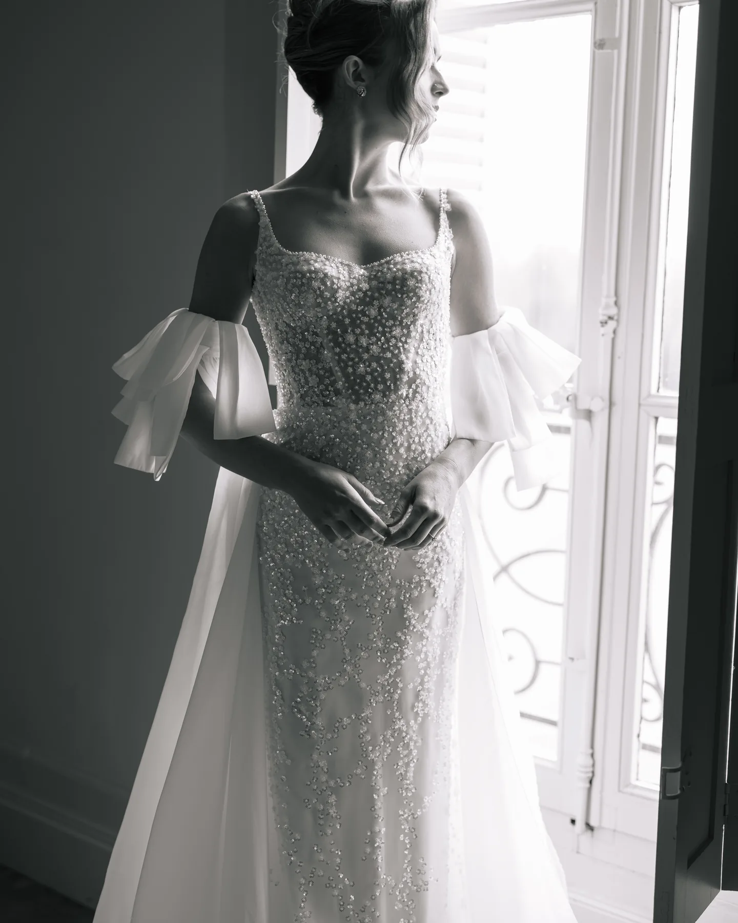 Mariée en robe pailletée devant une haute fenêtre, en noir et blanc