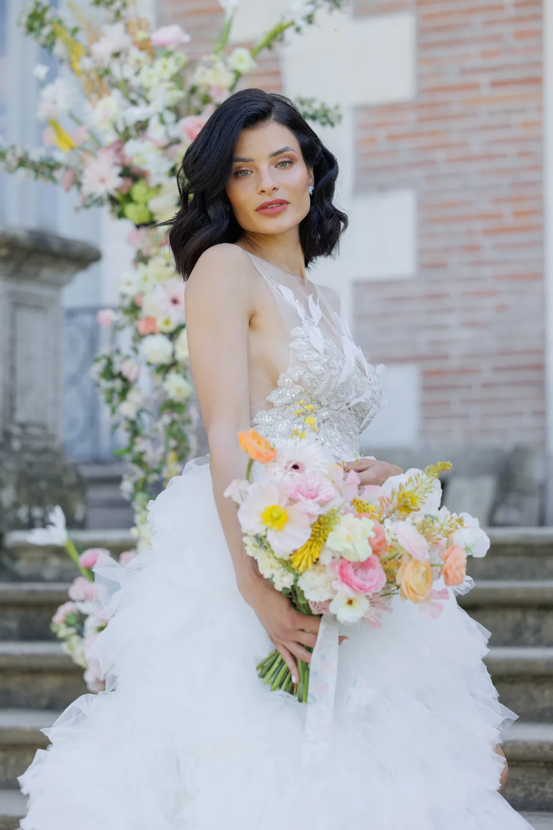 La mariée en robe de tulle tient un bouquet coloré devant une façade de brique